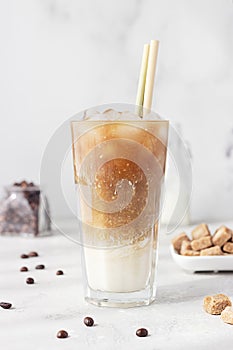 Cold coffee with milk and ice cubes in a tall glass with bamboo straw, light grey background.