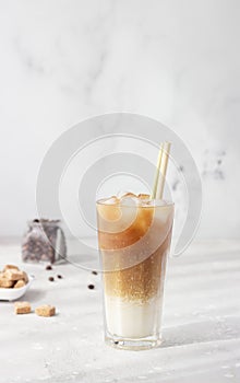 Cold coffee with milk and ice cubes in a tall glass with bamboo straw, light grey background.
