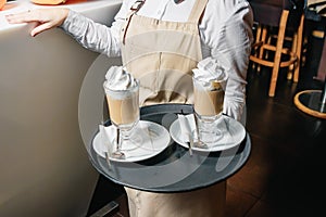 Cold coffee with whipped milk and caramel ice cream in high glasses on a tray in the hands of the waiter, selective focus