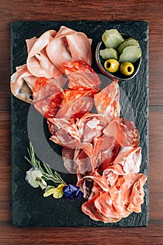 Cold cuts on stone plate with prosciutto, bacon, salami and sausages decorated wth flowers on wooden background.