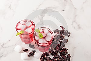 A cold drink made from hibiscus rose petals with ice cubes and mint on a marble table and dry rosebuds