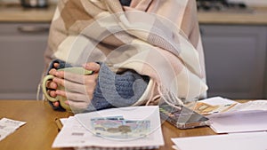 cold freeze woman warming up hands holding hot cup calculating money bills utilities checks at kitchen table