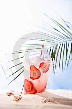 Cold fresh pink alcohol cocktail with ice, straw in plastic mug in sunlight on tropical beach with ocean view, vertical. Takeaway.