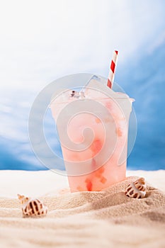 Cold fresh pink cocktail with ice in plastic mug on tropical beach, copy space, vertical. Vacation and picnic summer on ocean.