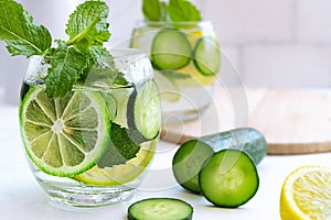Cold fresh summer drink in glass on bright grey brick wall background. Tasty lemonade with lemon, lime, cucumber, mint and ice.