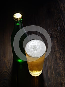 The cold frothy beer in a glass and a green bottle on a dark wooden background