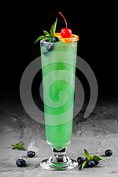 Cold green cocktail with blueberries, orange and ice in tall glass on dark background.