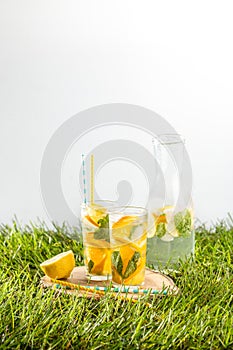 Cold lemonade drink with orange and mint in tall glass on green grass lawn wooden background