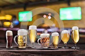 Cold mugs and glasses of beer on the old wooden table. Blurred pub interior at the background. Assortment of beer