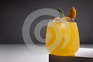 Cold orange fruit drink with ice, green rosemary, peach slice in glass on podium in elegant minimal dark white and grey abstract.