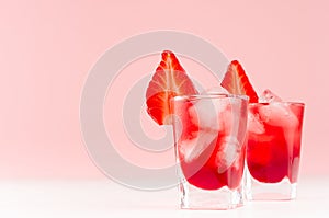 Cold red alcoholic liquors with ice cubes, strawberry slice in two shot glasses on white wood table and pastel pink wall.