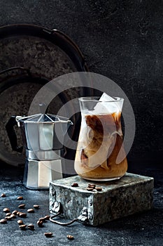 Cold refreshing iced coffee with cream in a tall glass and coffee beans on dark background.