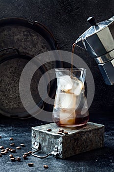 Cold refreshing iced coffee in a tall glass and coffee beans on dark background. Pouring coffee from moka pot into glass with ice