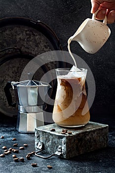 Cold refreshing iced coffee in a tall glass and coffee beans on dark background. Pouring cream into glass with iced coffee.