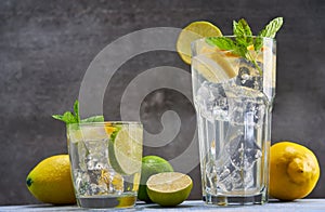 Cold refreshing limonade, mojito or gin tonic in glass, with fresh mint and ice cubes, lime and lemon on wooden board and black