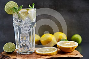 Cold refreshing limonade, mojito or gin tonic in glass, with fresh mint and ice cubes, lime and lemon on wooden board and black