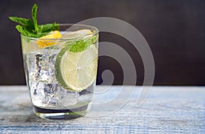 Cold refreshing summer limonade, mojito or gin tonic in glass, with fresh mint and ice cubes, lime and lemon on wooden table, on