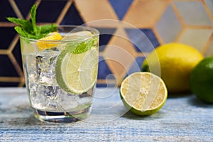 Cold refreshing summer limonade, mojito or gin tonic in glass, with fresh mint and ice cubes, lime and lemon on wooden table, on