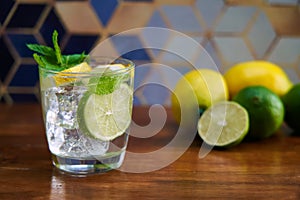 Cold refreshing summer limonade, mojito or gin tonic in glass, with fresh mint and ice cubes, lime and lemon on wooden table, on