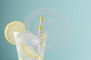 Cold shine sparkling refreshing drink for party with lemon slices, ice cubes, tonic, yellow straw in elegant glass on mint color.