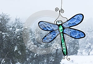 Cold snow day with oak trees seen through a window with bright blue and green stained-glass dragonfly sun catcher hanging in windo