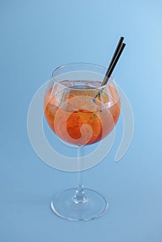 Cold summer alcohol cocktail with ice and orange in a wine glass and black plastic straw on light pastel blue background