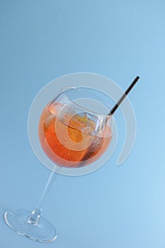 Cold summer alcohol cocktail with ice and orange in a wine glass and black plastic straw on light pastel blue background