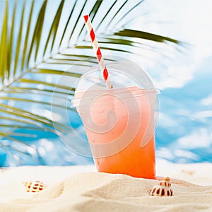 Cold tropical fruit cocktail in transparent plastic glass takeaway with straw on sunny coast with palm, sand, ocean view, square.