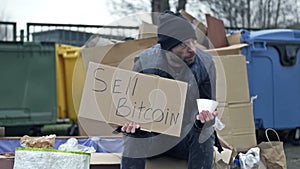Cold, unkempt-looking young man sits with a begging cup by a pile of rubbish and holds up a handwritten SELL BITCOIN