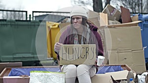 Cold, unkempt-looking young woman sits with a begging cup by a pile of rubbish and holds up a handwritten HUNGRY poster