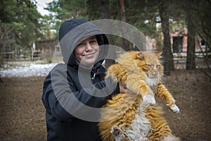 On a cold winter day, a teenage boy sits on a bench with a ginger cat in the forest