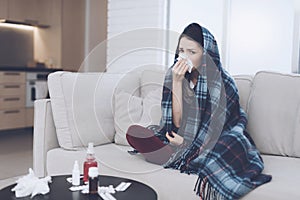 A cold woman sits on a light couch wrapped in a blue checkered whip. She blows her nose into a napkin