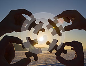Collaborate four hands trying to connect a puzzle piece with a sunset background.