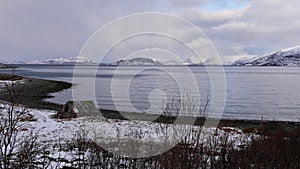 Collapsed old wooden shed at the shore of Altafjord, Norway with bare trees and snow-covered mountains on cloudy day in winter.