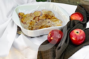 Collapsed soft baked apples in a white ceramic square shape on a special package. A white tablecloth is laid carelessly on the tab