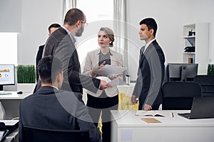 Colleages stand around in the office, woman looks at her chief and listens