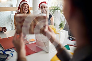 Colleague showing wrapped present to two female coworkers at the office with santa clause hats on their hads