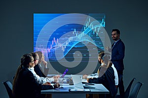 Colleagues in corporate environment gather in boardroom, studying digital stock chart displayed on monitor during