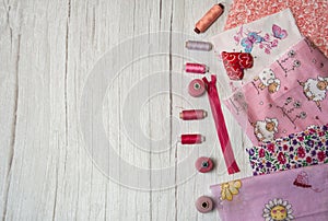 A collection of fabrics in different shades of pink, reels of thread and pins on a light wooden background.