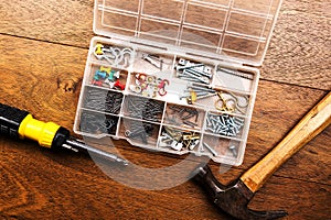 Collection of nails, thumb nails, screws, hooks, pins, hanger, plastic and bolts in plastic box next to a hammer and a screwdriver