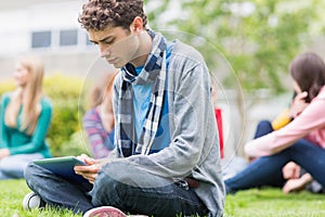 College boy using tablet PC with blurred students in park