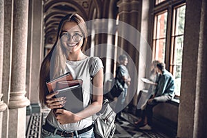 College life.Beautiful student in glasses holding books and looking at camera