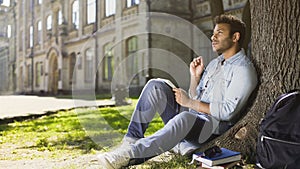 College student sitting with notebook under tree doing home assignment, studying