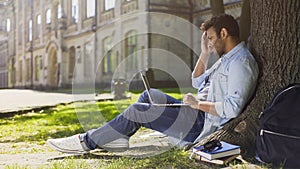 College student sitting under tree with laptop looking upset disappointing news photo