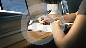 College or university student doing school homework at home. Working late at night. Young woman writing on paper with pen.