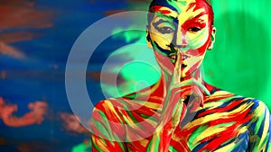 Color face art and body paint on woman showing silence sign the finger near lips. Abstract portrait of the bright