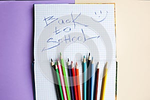 Color pencils lying on pastel purple and beige background. Squared notebook with back to school concept sign and smile. Colorful