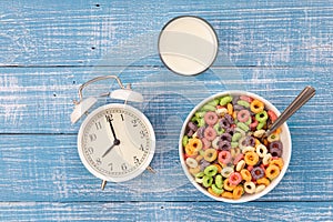 Colored breakfast cereals, alarm clock and glass of milk on a wooden background.