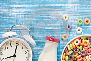 Colored breakfast cereals in a bowl, alarm clock and bottle of milk, close up.