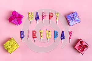 Colored candles on cake in the form of letters happy birthday, gift boxes on pink background